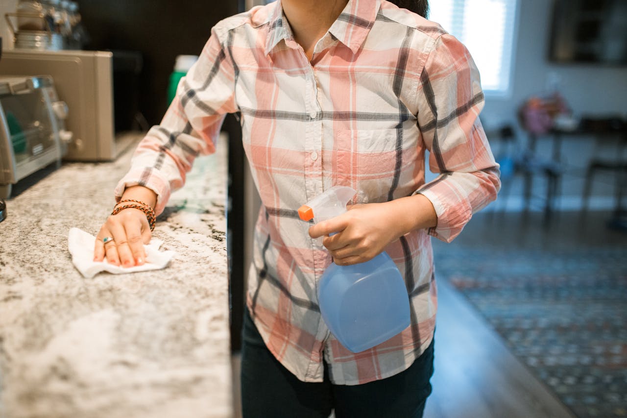about-01 A woman wearing a plaid shirt cleans a kitchen counter using a spray bottle and cloth.