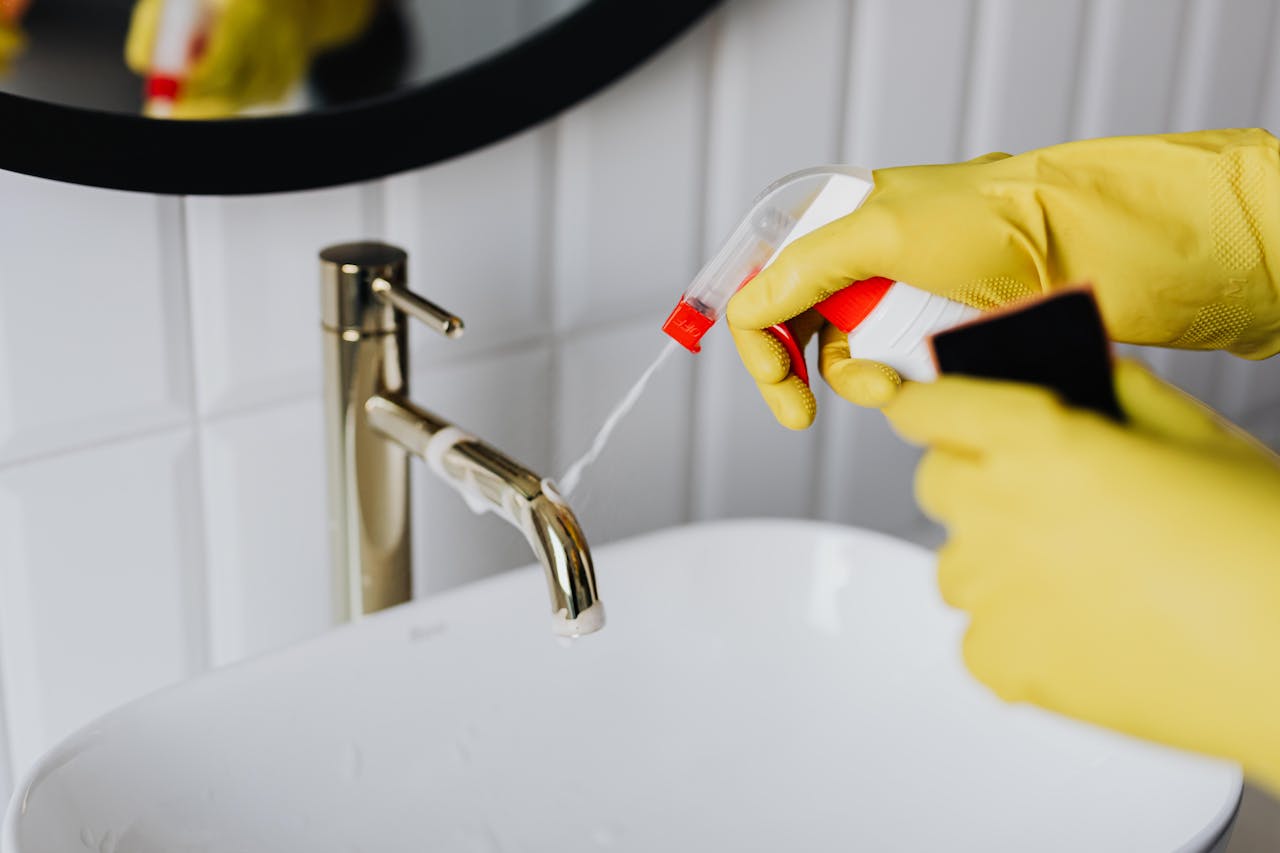 services-01 From above anonymous housekeeper in yellow rubber gloves spraying cleaner to chrome water faucet in bathroom